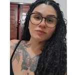 Mulher branca com tranças, oculos de grau com armação preta arredondada, tatuagem no peito e ombro com vestido preto.